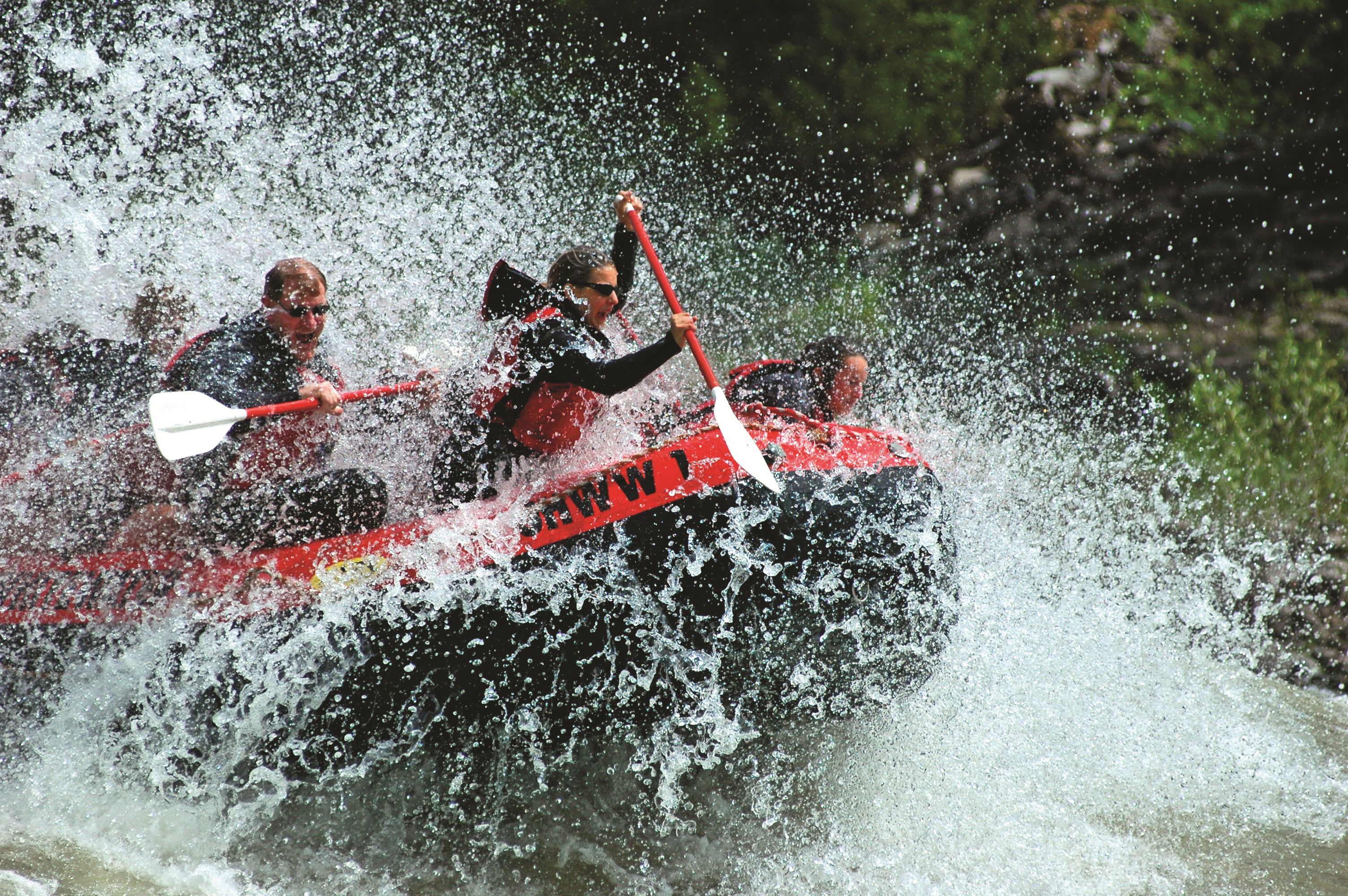 Un groupe de personnes à bord d’un bateau rouge et noir navigue sur une rivière, l’eau éclaboussant tout autour.