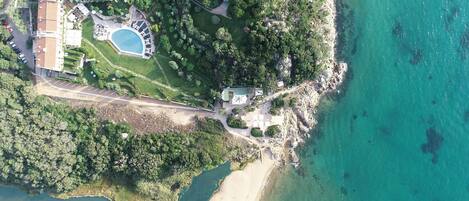 An aerial view of a coastal property with a swimming pool, landscaped gardens, and a beach.