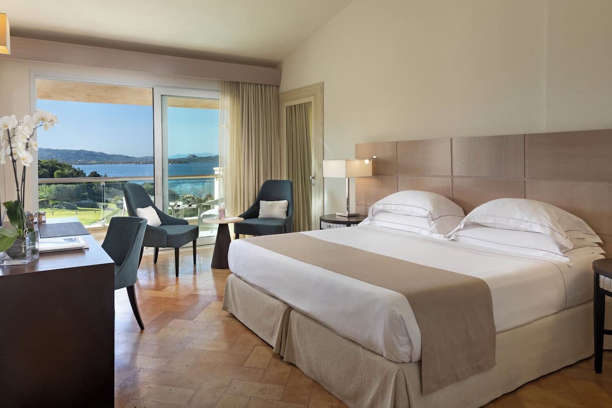 A hotel room with a large bed, two chairs, a desk, a lamp, and a view of the sea.