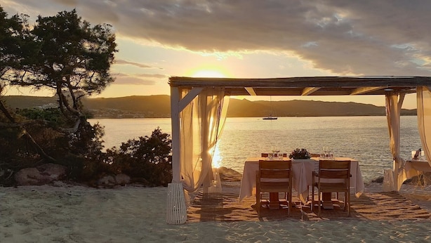 A beachside dining setup with a table and chairs under a canopy, overlooking a calm sea and distant mountains. - L'Ea Bianca Luxury Resort (Arzachena)