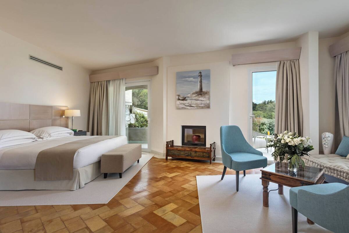 A hotel room with a bed, two armchairs, a coffee table, a fireplace, and a painting of a lighthouse.