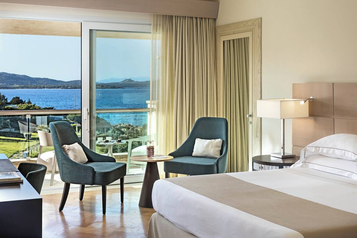 A hotel room with a bed, two armchairs, a small table, a lamp, and a view of the sea.
