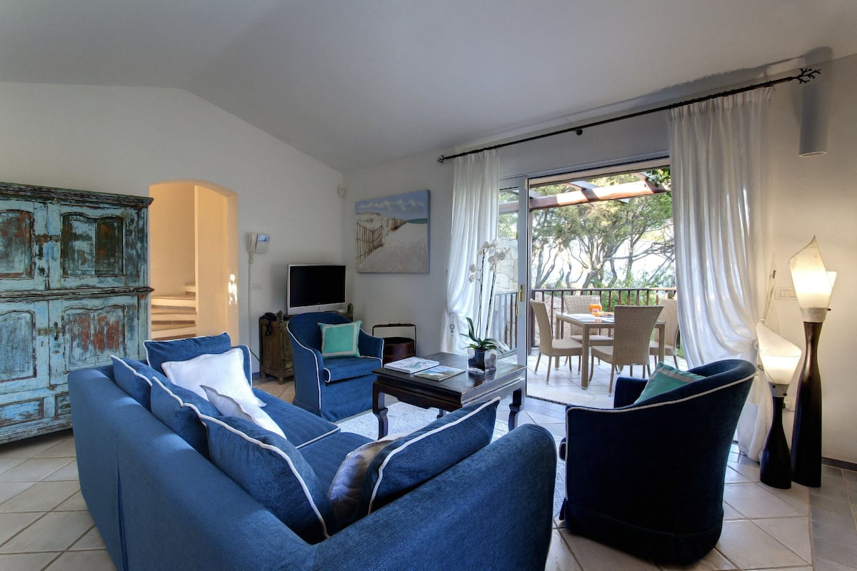 A living room with a sofa, armchairs, a coffee table, and a television. There is a dining area with a table and chairs outside.