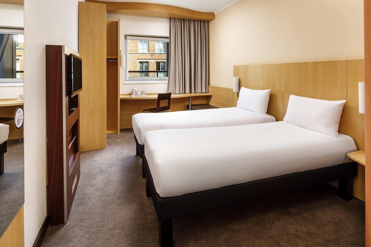 A hotel room with two beds, a desk, a chair, a TV, and a window with curtains.