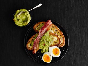 A plate with toasted bread, two halves of a boiled egg, and a piece of bacon, accompanied by a bowl of green spread.