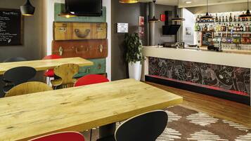A restaurant interior with wooden tables, black and red chairs, hanging pendant lights, a bar area with bottles, and a menu board.