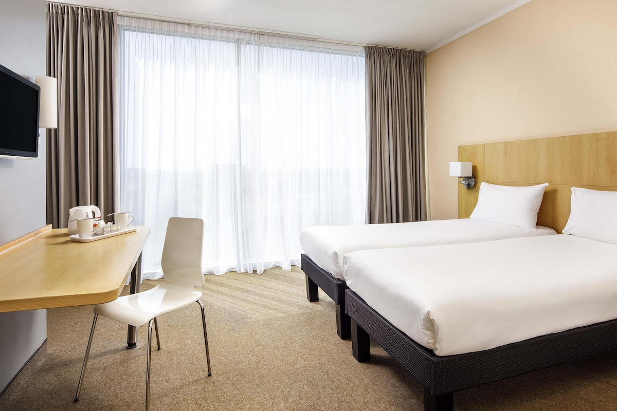 A hotel room with a bed, a desk with a chair, a TV, and a window with curtains.