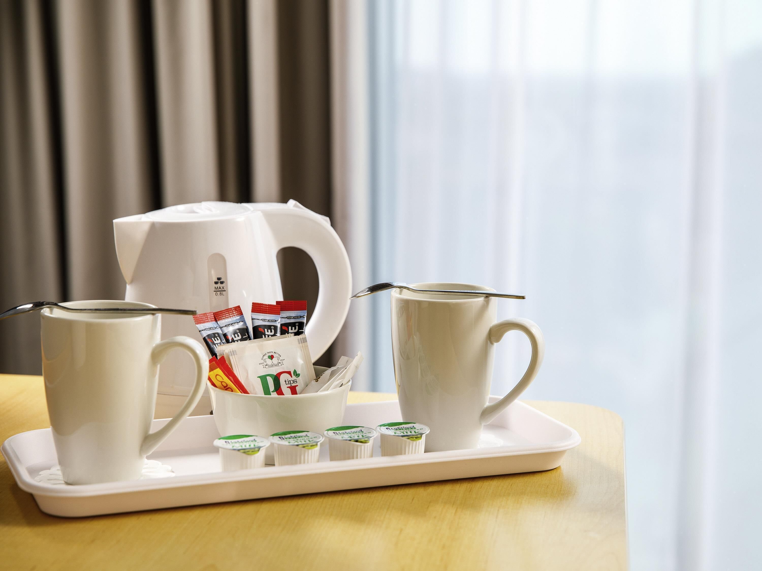 Una bandeja con dos tazas blancas, una tetera blanca y sobres de café instantáneo sobre una mesa.