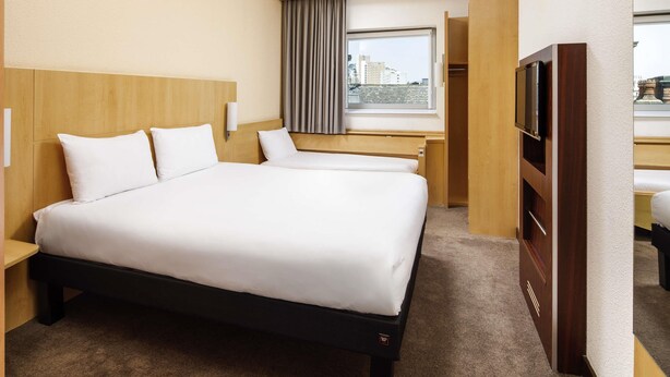 A hotel room with a large bed, a desk, a television, and a window with a city view. - ibis Nottingham Centre (Nottingham)