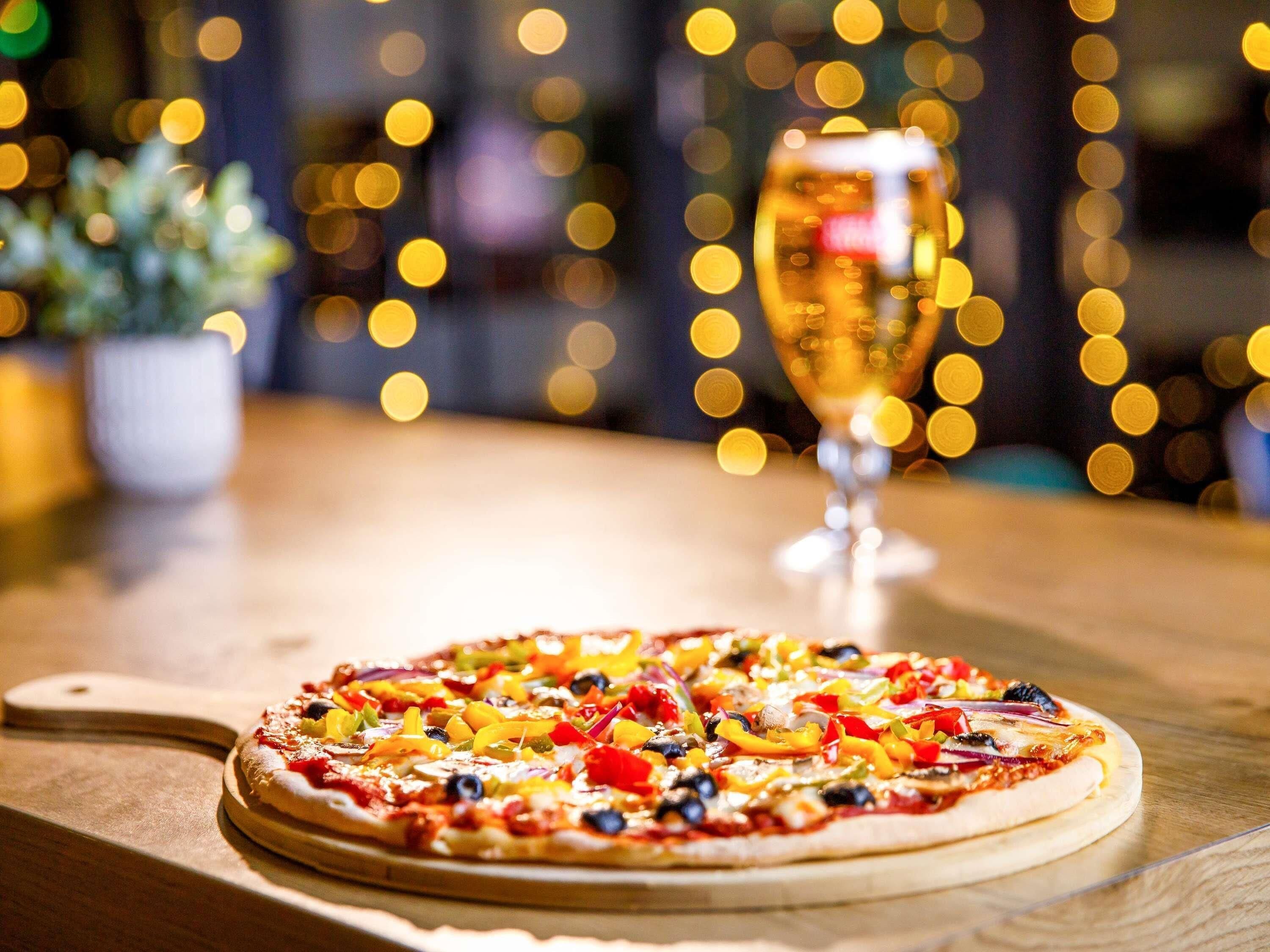 A pizza on a wooden board with a glass of wine in the background.