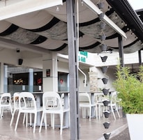 Uma área de estar ao ar livre com cadeiras e mesas de plástico branco, um arranjo de plantas pendente e um edifício com uma placa que diz "Restaurant & Grill".