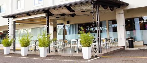 Un área de café al aire libre con mesas, sillas, plantas en macetas y un espacio de estar cubierto.