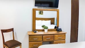 Ein Hotelzimmer mit einem Holztisch, einem Sessel, einem Bett mit weißer Bettwäsche, einer kleinen Topfpflanze, einem Telefon und einem an der Wand befestigten Fernseher.