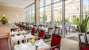 Un restaurante con amplios ventanales, mesas dispuestas con manteles blancos, sillas rojas y plantas en macetas.