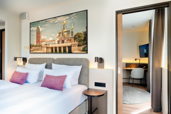 Una habitación de hotel con cama, almohadas, una mesita, un escritorio con silla y un cuadro de un edificio en la pared.
