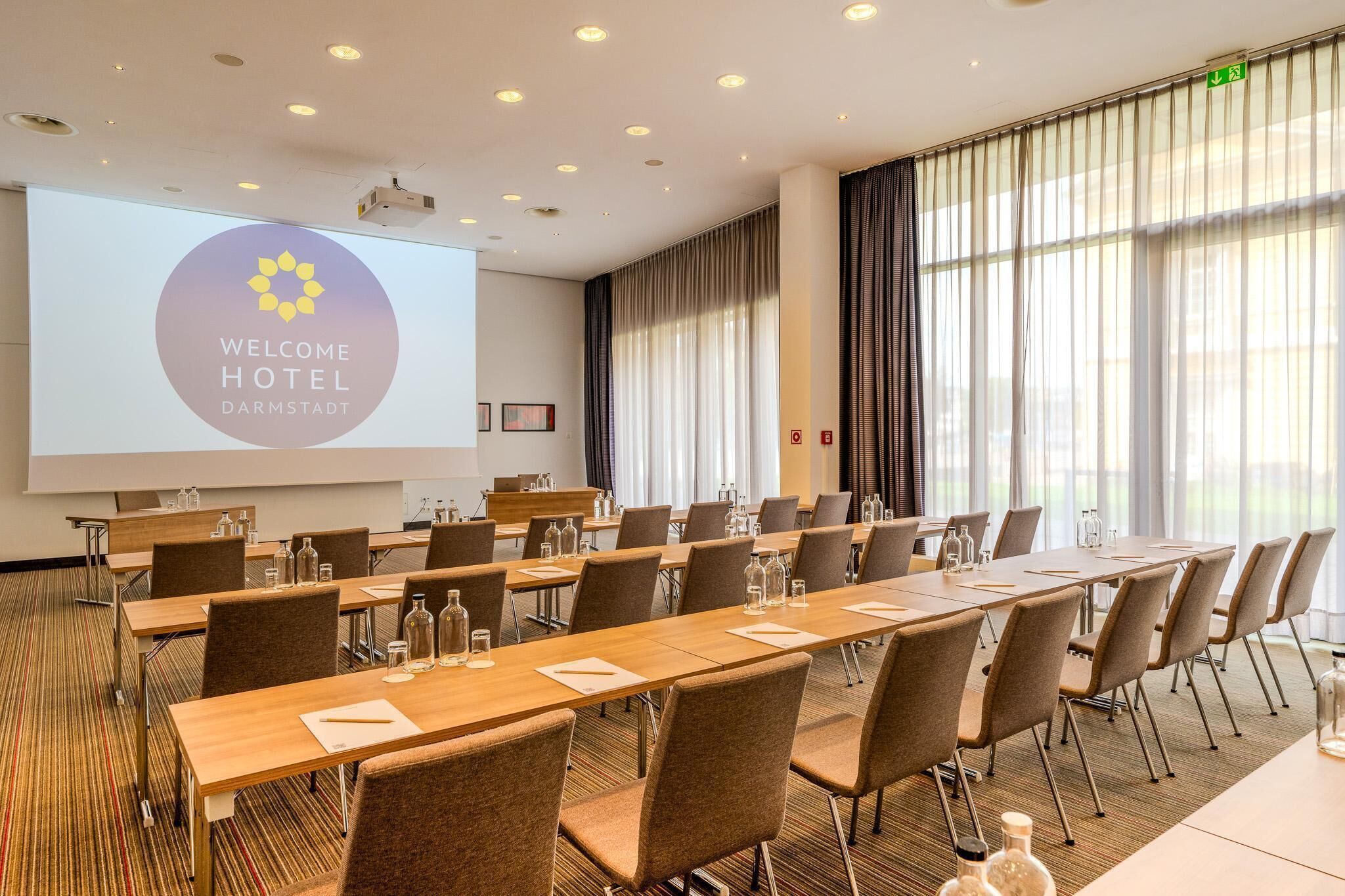 Un salón de conferencias con una larga mesa, sillas, una pantalla de proyector que muestra “WELCOME HOTEL DARMSTADT” y grandes ventanales con cortinas.