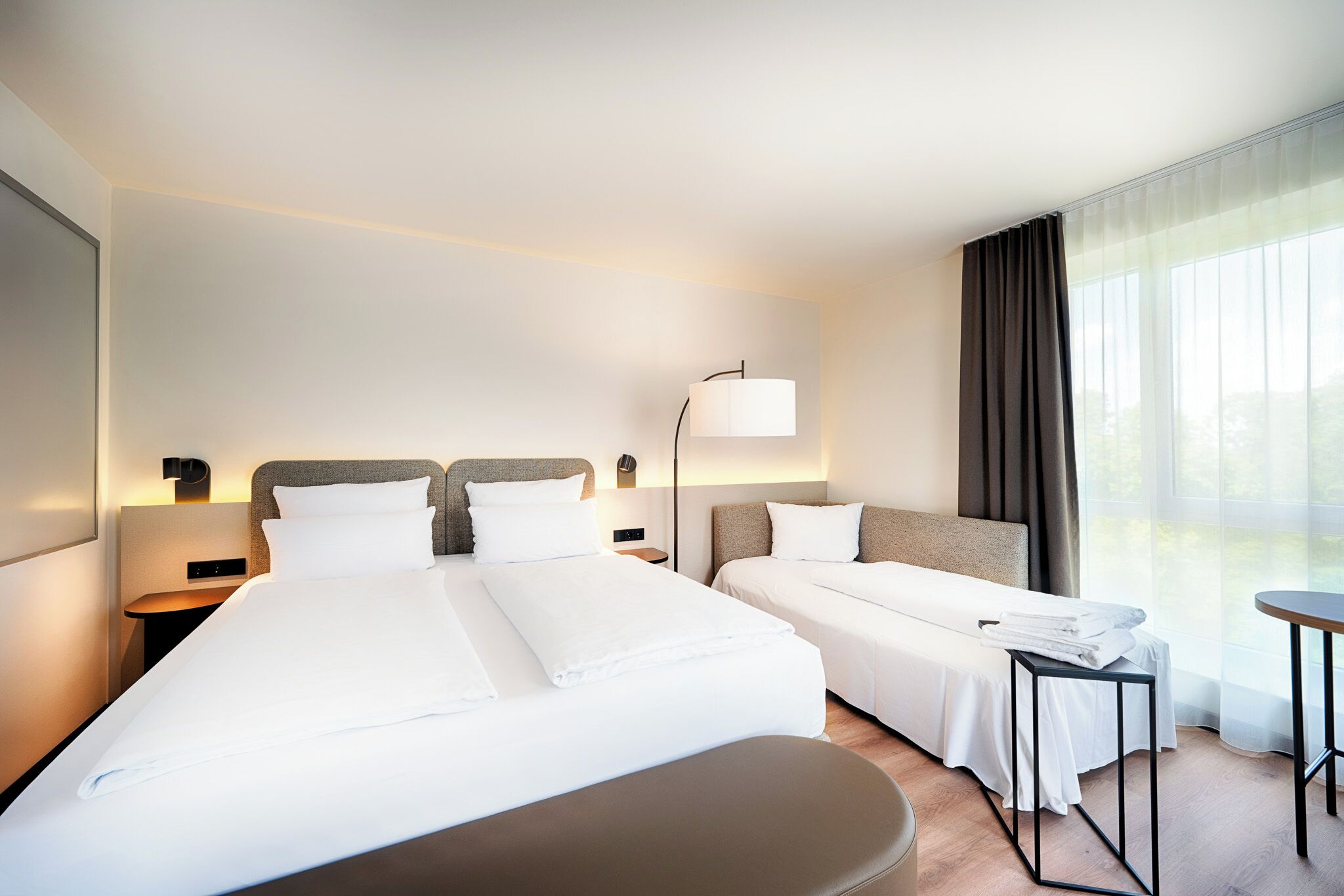 A hotel room with two beds, a bedside table with a lamp, a small table, and a large window with curtains.