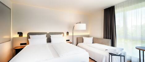 A hotel room with two beds, a bedside table with a lamp, a small table, and a large window with curtains.