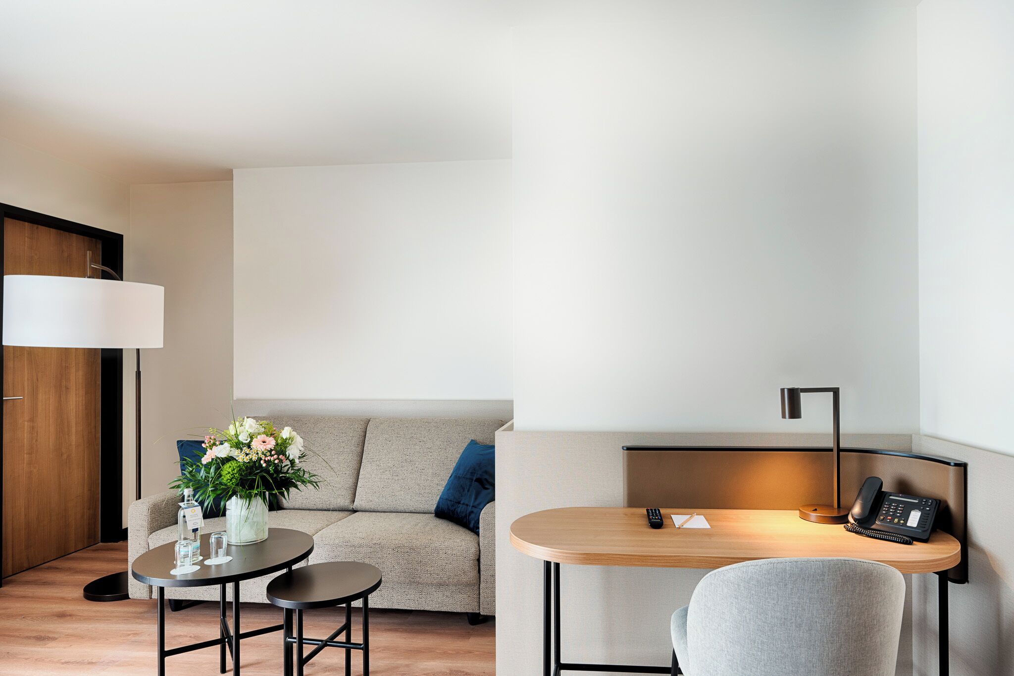 Ein modernes Hotelzimmer mit einer Couch, einem kleinen runden Tisch mit einer Lampe, einem Schreibtisch mit Stuhl und einem Telefon.