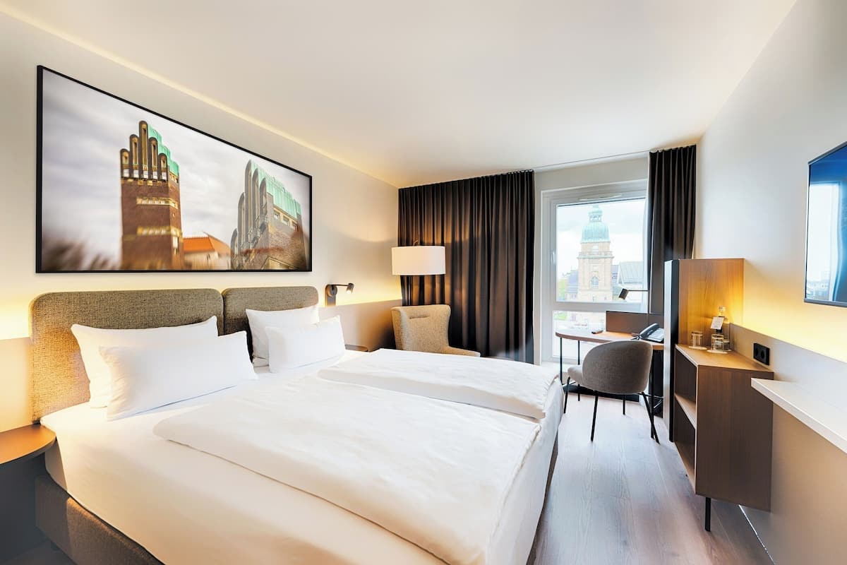 A hotel room with a large bed, a desk, a chair, a lamp, a TV, and a cityscape view.