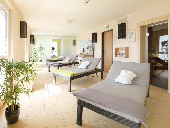 A hotel room with two reclining beds, a potted plant, a reception desk, and a bathroom visible through a glass door.
