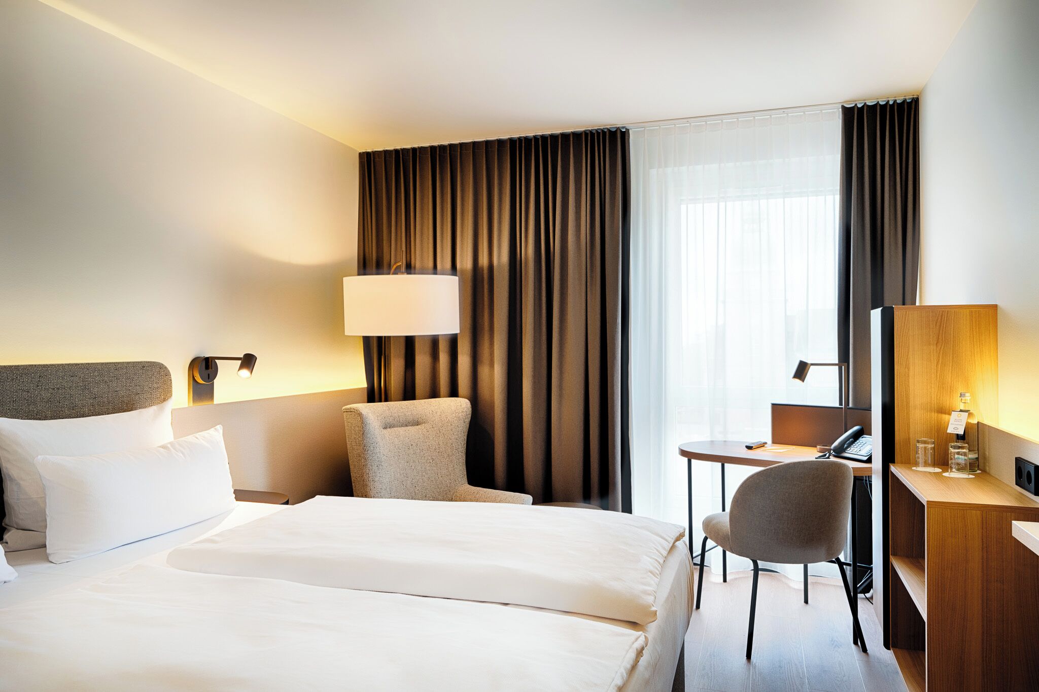 Ein Hotelzimmer mit einem Bett, einem Schreibtisch, einem Stuhl, einer Lampe und einem Fenster mit Vorhängen.