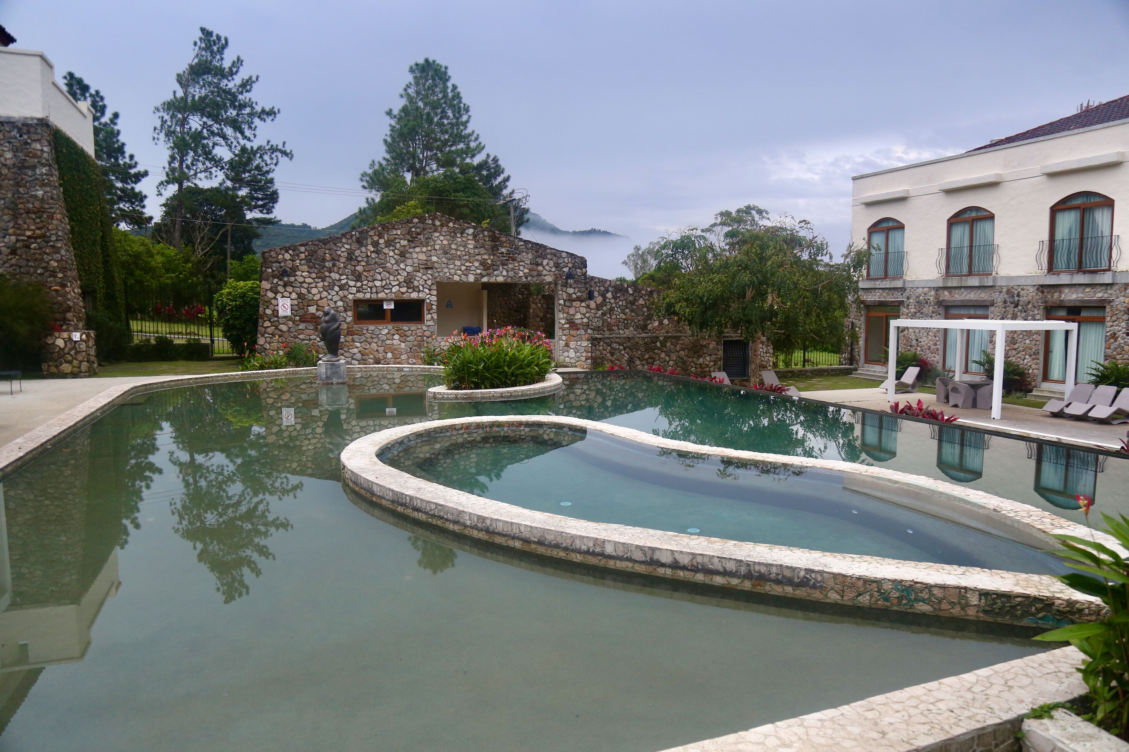 A swimming pool with a stone border, surrounded by a stone building and a white house with a balcony.