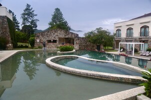 Una piscina con borde de piedra, rodeada por un edificio de piedra y una casa blanca con balcón.