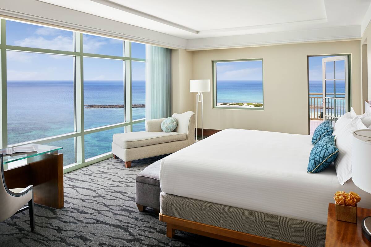 Camera d'albergo con un'ampia finestra vista mare, un letto con testiera a motivo a spina di pesce, una scrivania con piano in vetro e una lampada su un comodino.
