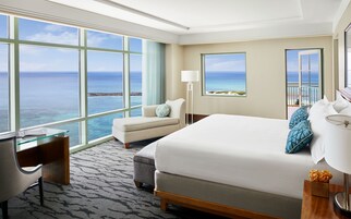 Habitación de hotel con un gran ventanal que ofrece vistas al mar, una cama con cabecera de patrón espiga, un escritorio con superficie de vidrio y una lámpara en la mesita de noche.