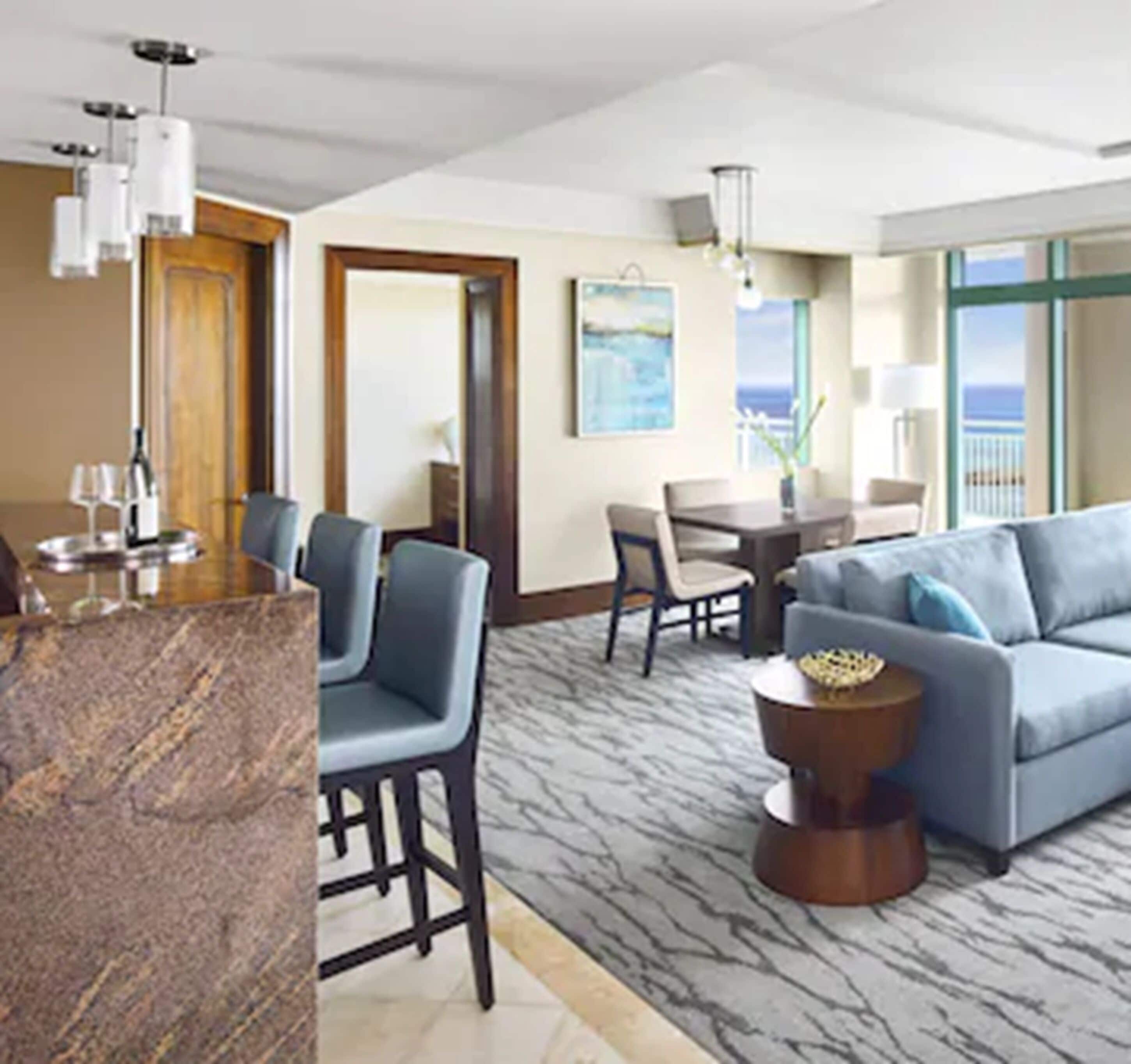 A modern hotel room with a marble bar, a seating area with a sofa and chairs, a dining table, and a view of the ocean through large windows.