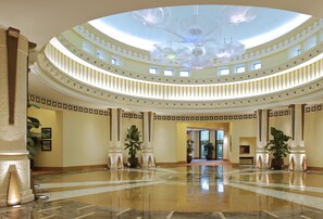 A grand, circular lobby with a high ceiling and a central chandelier. The lobby features a polished marble floor and is adorned with large potted plants.