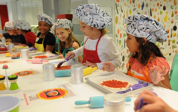 A group of people wearing chef hats, engaged in a cooking activity with pizza dough and sauce on a table.
