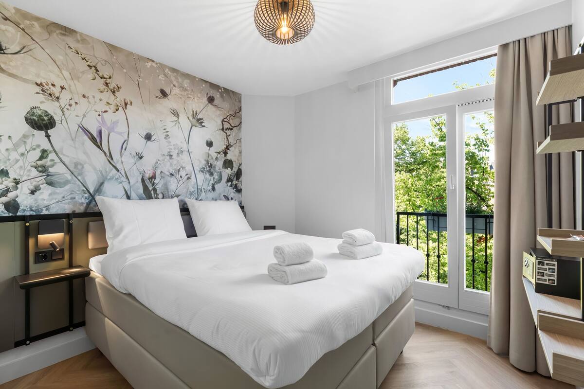 A modern hotel room with a large bed, a mural on the wall, a balcony with a view, and a built-in desk with a lamp.