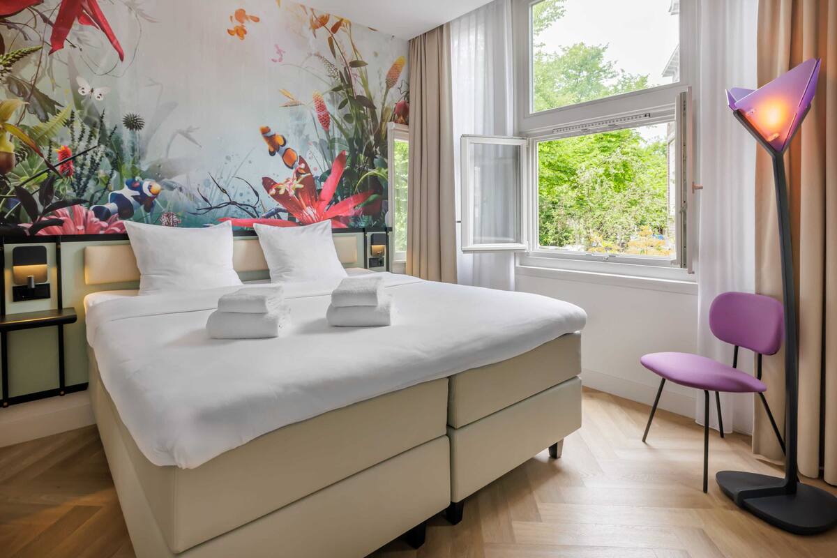 A bedroom with a bed, a chair, a nightstand, a lamp, and a window with a view of greenery.