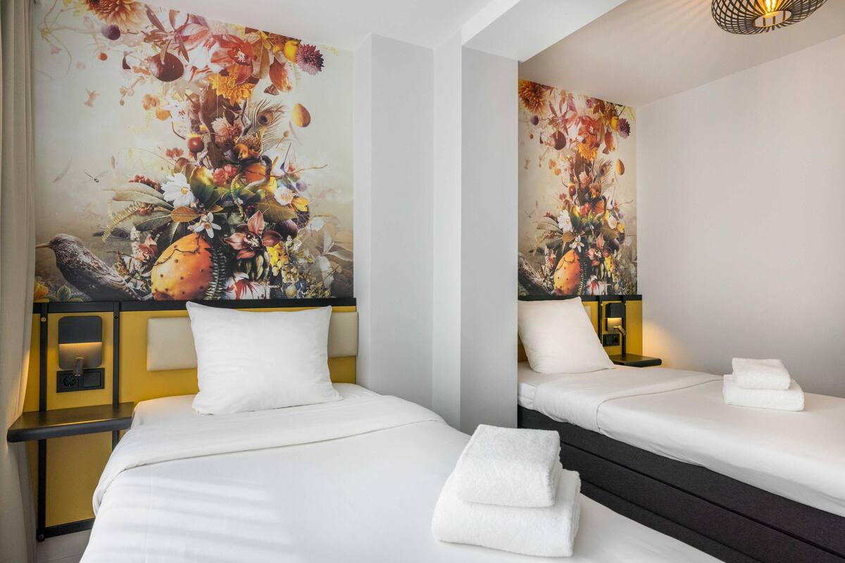 A hotel room with two beds, white bedding, a large floral artwork on the wall, and a bedside table with a lamp.