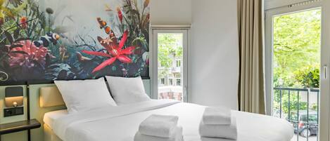 A hotel room with a large bed, two pillows, a nightstand, a painting, a window with a view of greenery, and a balcony.