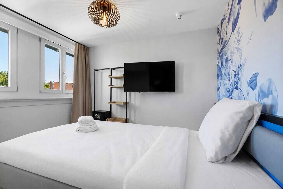 A hotel room with a bed, a flat-screen TV, a wall-mounted shelf, and a window with curtains.