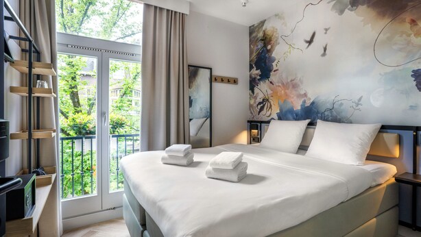 A modern bedroom with a large bed, a TV, a balcony with curtains, and a floral mural on the wall. - Dutch Design Hotel Vondelpark (Amsterdam)
