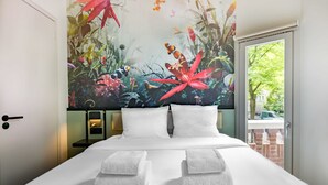 A hotel room with a bed, white towels, a nightstand, a wall mural, and a window with a view of greenery. - Dutch Design Hotel Vondelpark (Amsterdam)