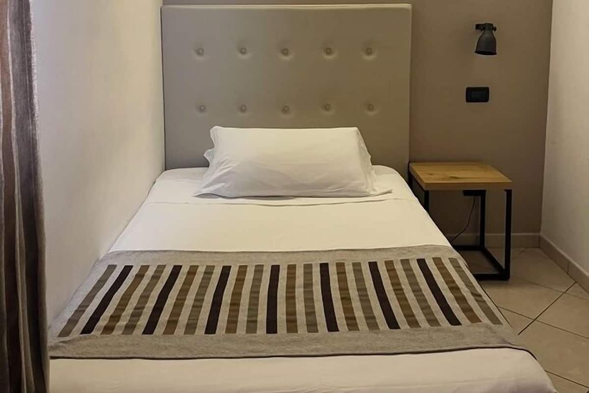 A compact hotel room with a single bed, a small bedside table, and a wall-mounted light fixture.