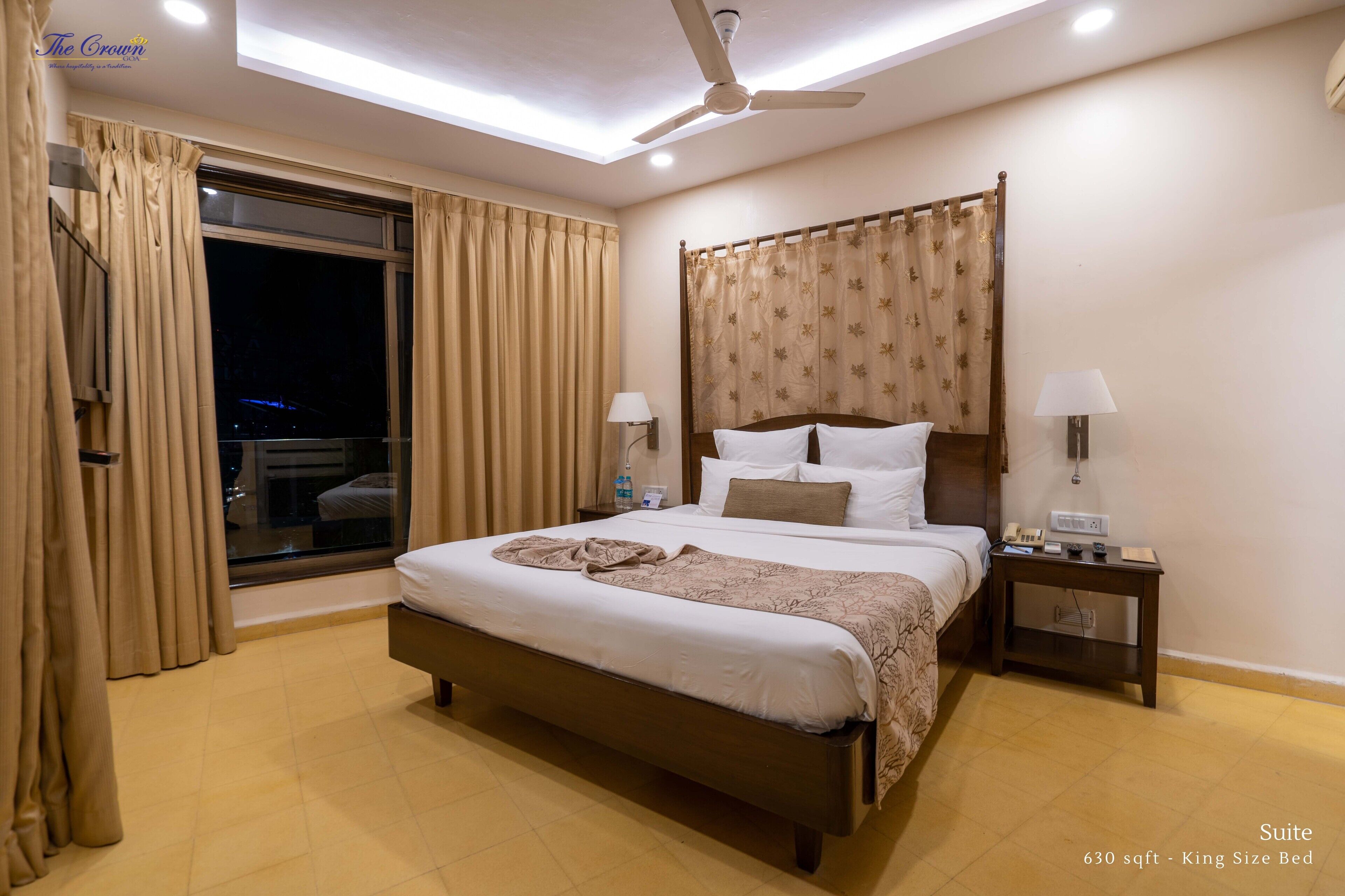 Eine Hotel Suite mit einem Kingsize-Bett, Deckenventilator, Nachttisch mit Lampe und Blick durch ein mit Vorhängen drapiertes Fenster.