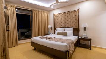 Eine Hotel Suite mit einem Kingsize-Bett, Deckenventilator, Nachttisch mit Lampe und Blick durch ein mit Vorhängen drapiertes Fenster.
