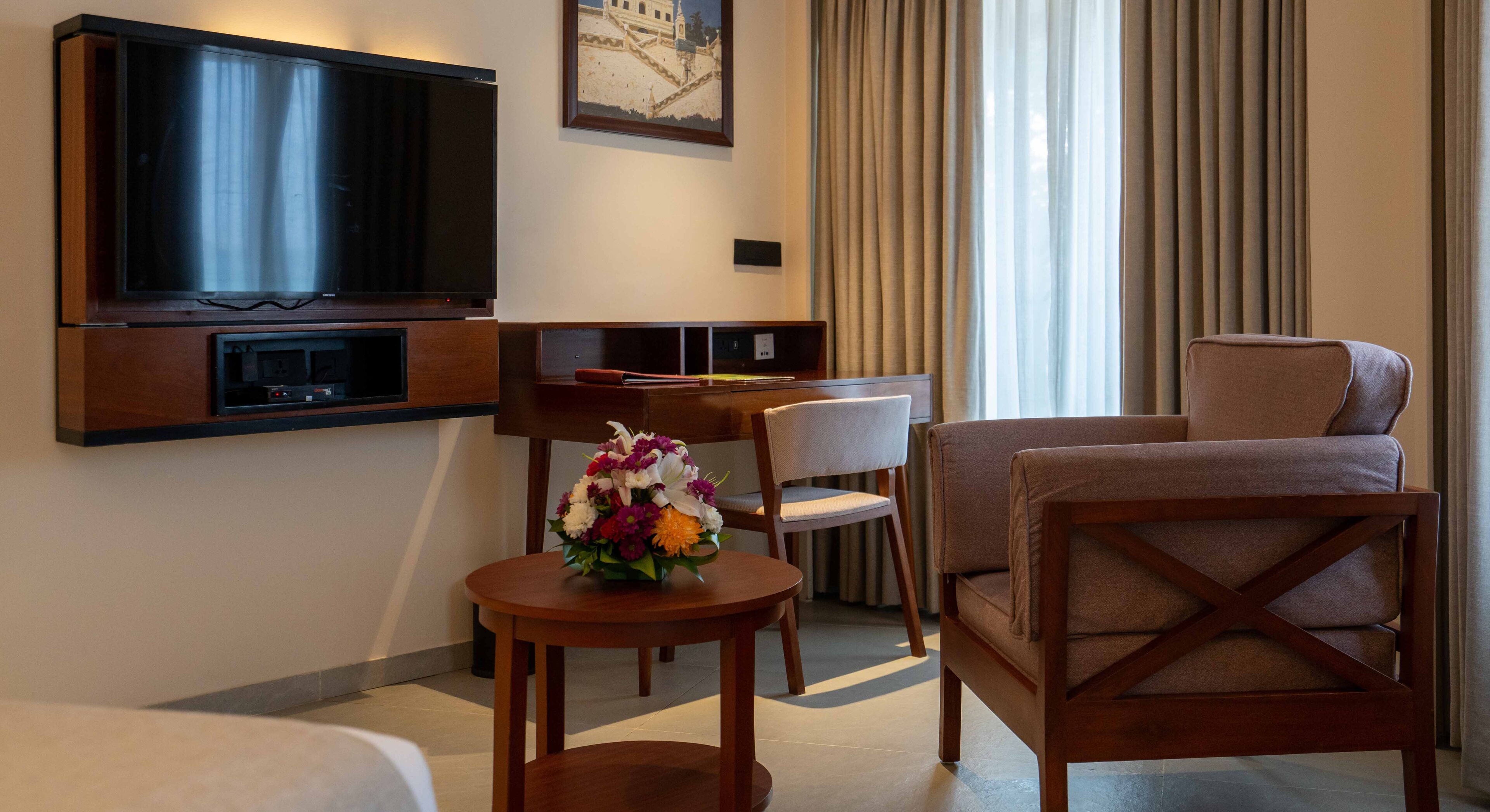 A hotel room with a TV, a desk, a chair, a sofa, and a small table with a flower arrangement.