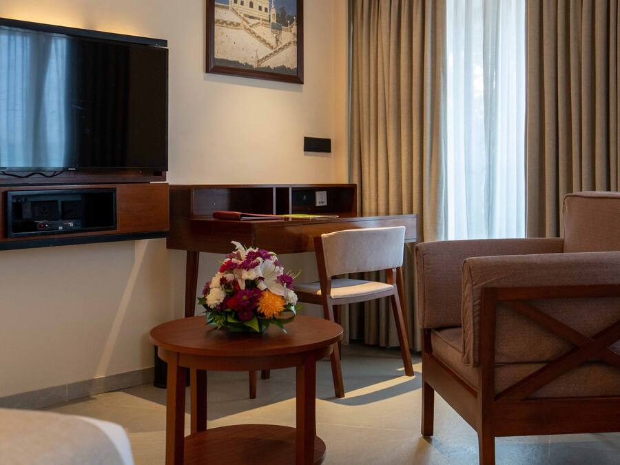 A hotel room with a TV, a desk, a chair, a sofa, and a small table with a flower arrangement.