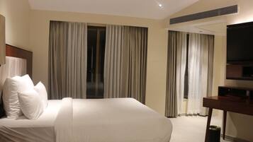 A hotel room with a large bed, a desk with a flat-screen TV, and two windows with curtains.