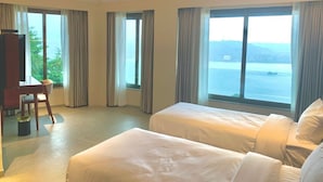 Ein Hotelzimmer mit zwei Betten, einem Schreibtisch und einem Sessel. Große Fenster mit Vorhängen bieten Blick auf das Meer.