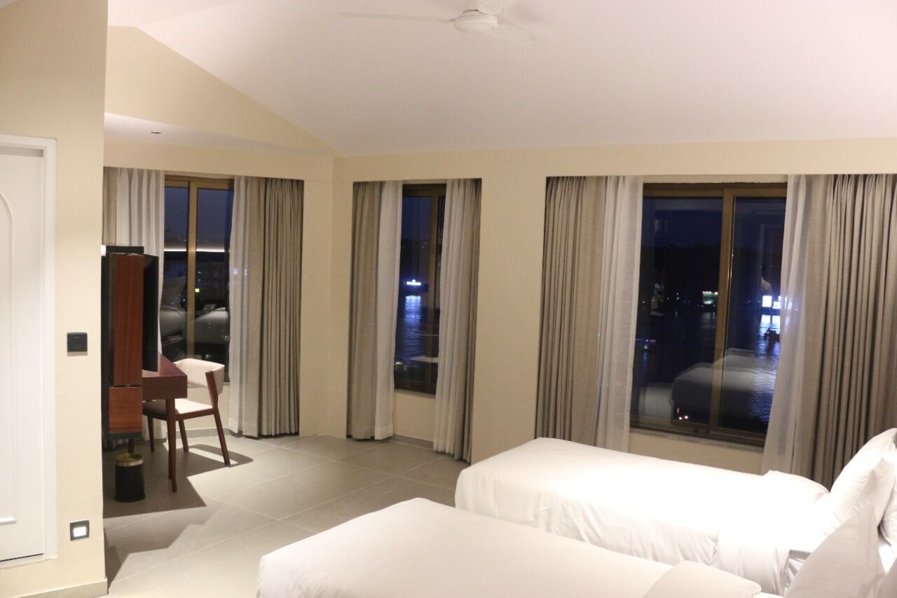 A hotel room with two beds, a desk, and a chair. There are large windows with curtains, and a view of the city at night.