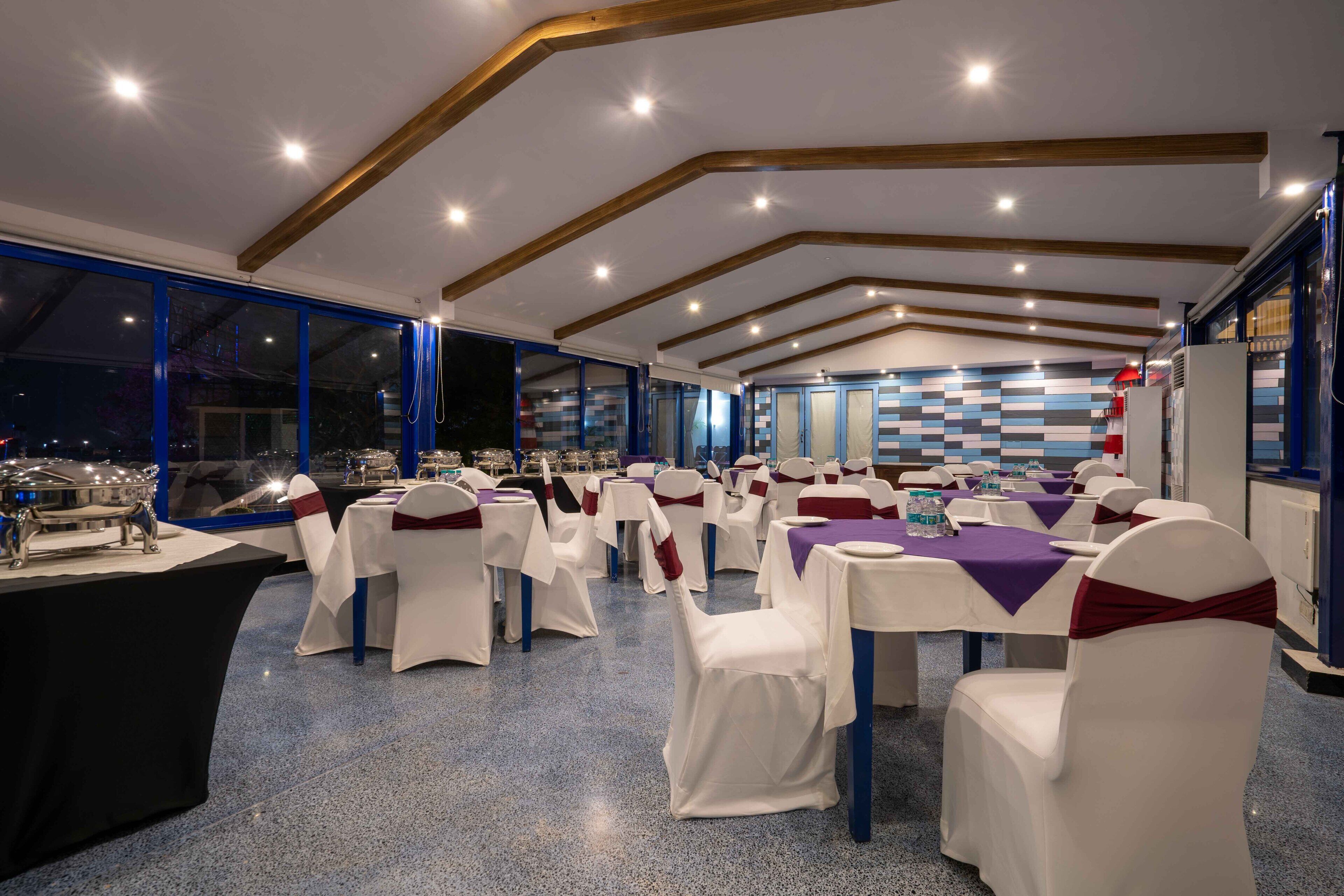 Une salle de banquet où les tables sont dressées pour un événement formel, avec un plafond au design moderne et de grandes fenêtres.