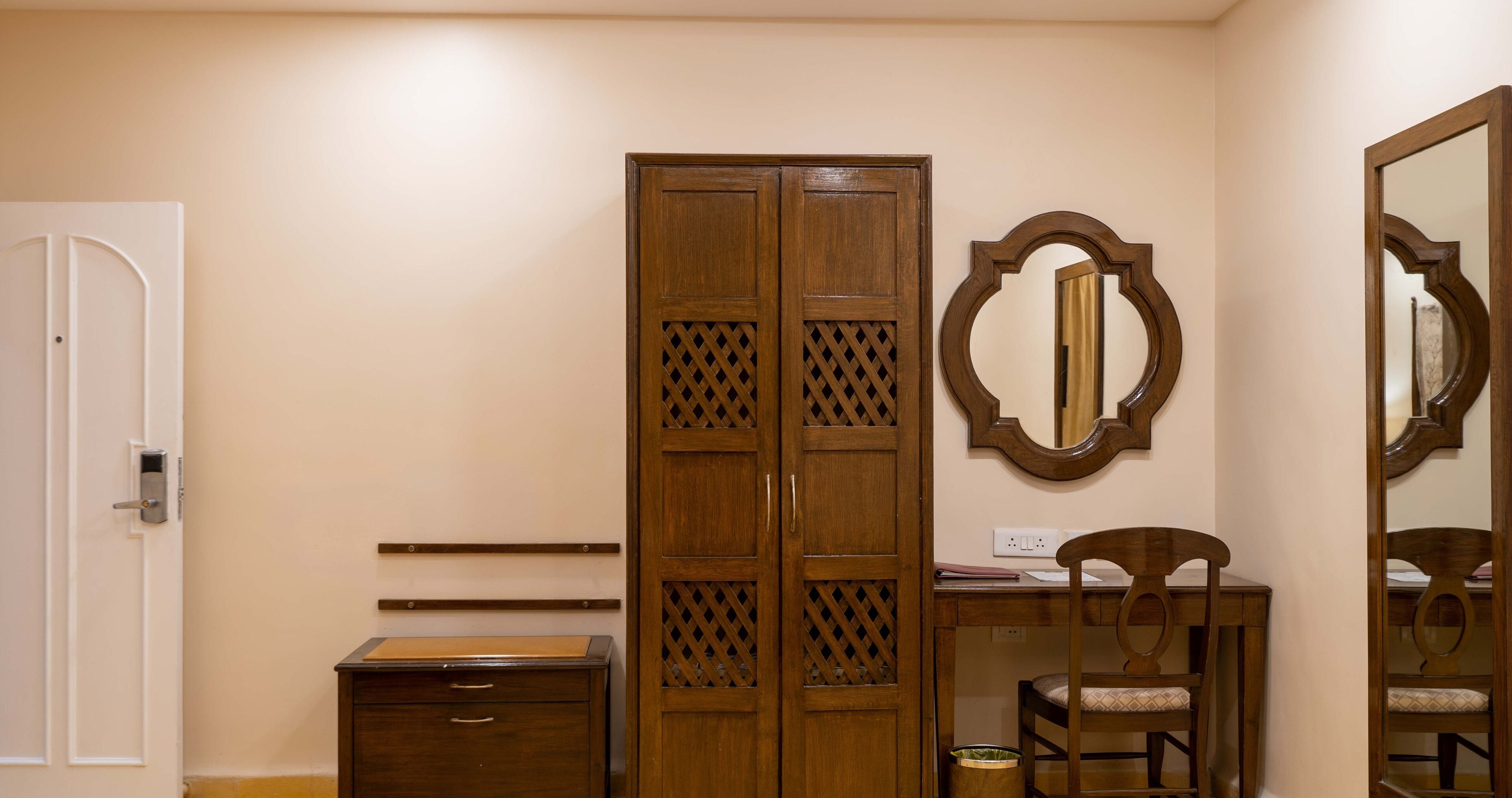 A room with a wooden wardrobe, a desk with a chair, and a mirror on the wall.
